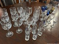 Photo showing assortment of clear and colored glass drinkware on table, including stemmed glasses, tumblers, and cocktail glasses
