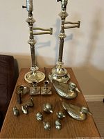Photo showing two tall lamp bases, brass duck and swan figurines, and candlesticks arranged on wooden surface