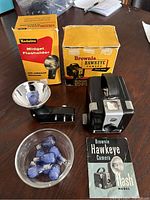 Brownie Hawkeye camera with original box, instruction booklet, Kodalite Midget Flashholder with reflector, and 8 bulbs on a table.