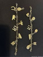 Photo showing two gold tone charm bracelets with various charms such as letters, a crown, rose, horseshoe, handbag, shoe, typewriter and other decorative charms against a black background.