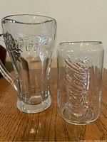 Side view of two embossed clear Coca-Cola glasses, one tall with handle, one shorter without.