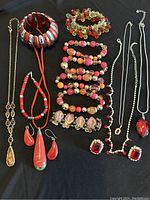 Photo showing assortment of red jewelry including chunky bracelet, multiple beaded bracelets, necklaces with pendants and matching earrings and ring on black surface.