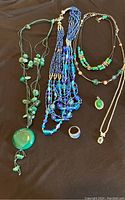 Full view of all six jewelry items including four necklaces, pendant, and ring in blue and green tones laid out on brown background.