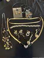 Photo of assorted gold and black jewelry including necklaces with pendants and beads, bracelets in gold-tone, and multiple pairs of earrings with black and amber stones.