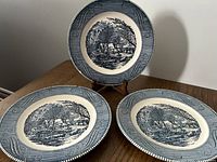 Three blue and white underglaze porcelain plates displayed on a wooden table, each featuring a print of 'The Old Grist Mill' by Currier & Ives with decorative borders.