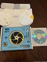 Open box showing unassembled blue engine base with brass ring, black flywheel, and additional small assembly parts inside blue foam. Instruction CD is beside the box.