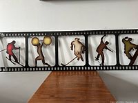 Front view of black metal film strip wall hanging with sports silhouettes showing skier, weightlifter, hockey player, baseball and basketball players.