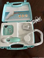 Top view of open case showing handheld device, control panel with SCAN and SINGLE buttons, round docking station, and power adapter, all arranged inside light blue hard plastic carrying case with handle.