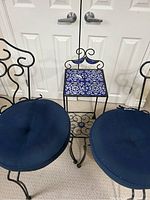 Front view of two black rod iron chairs with blue cushions and black rod iron table with blue and white tile top between them.