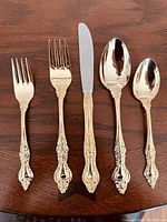 Close-up of five flatware pieces: salad fork, dinner fork, dinner knife, soup spoon, and teaspoon, showing ornate gold plated handles with floral scroll design.