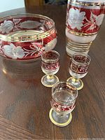 Front view showing bowl, vase, and three shot glasses on table highlighting red and gold colors with etched floral designs.