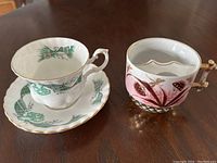 Photo showing the two vintage cups on a wooden table - a white tea cup and saucer with green plaid floral pattern, and a pink mustache mug with a unique rim shape and gold handle.
