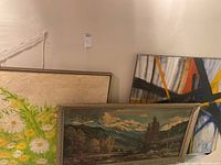 Three art pieces leaning against wall, including floral canvas in front, framed landscape print, and unframed abstract painting behind