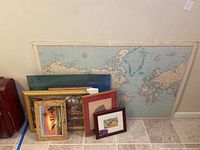 Wide view of large vintage map on poster board with assorted framed artworks leaning against the wall, showing overall lot contents and sizes