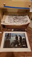 Various art prints and posters stacked under a small table, including visible black and white character posters and a color fishing scene print laid on the floor.