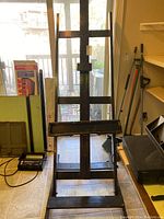 Full front view of the vintage wooden artist's easel standing opened on a tiled floor with background items visible, showing height and structural details.