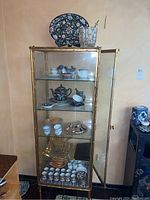 Full view of glass cabinet with various tea ware, mugs, dishes, bowls, and flatware inside and on top including decorative plate and ice bucket.