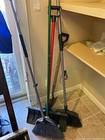 Photo showing two shovels and three brooms standing upright against a wall on tile floor.