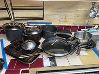 Wide shot showing assorted cookware including pots, pans, baking trays, and steamer insert placed on a kitchen counter.