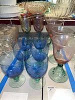 View of all goblets arranged with two blue bowl green stem goblets in front, other clear and purple tinted goblets behind, showing variety of sets.