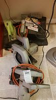 Photo showing multiple power tools including green sander, Black & Decker circular saw, and other electric tools stacked on tiled floor.