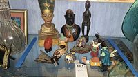 Photo of figurines and decorative objects arranged together including Egyptian bust, African statues, angels, red egg, and glass apple compact