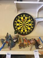Wide view of dartboard with darts and various carved wooden toys and game pieces including a small wooden wagon, spherical rattle, and picker sticks on shelf
