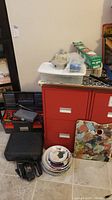 Overall view of the lot showing cordless drill in charger, tool cases, paint palettes, stacked plates, a red metal filing cabinet, and a box with plumbing supplies on tiled floor.