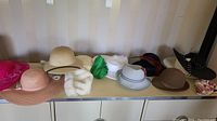 Wide shot of all 9 ladies hats arranged on a table showing a variety of colors, styles and materials including straw, fabric, faux fur and felt