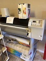 Front view of HP DesignJet 2500CP printer on stand with HP ink boxes on top and storage shelf below