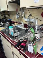 Kitchen counter with assorted kitchenware items including utensils, cooking thermometers, hand soap dispenser, and small gadgets in a sink and on countertop.