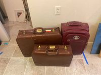 Three pieces of luggage: one burgundy Kathy Van Zeeland rolling suitcase with crocodile pattern and two vintage brown leather hard side suitcases with locks.