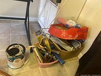 Photo of assorted hand tools in a pink container along with a paint can and a surge protector on a tiled floor
