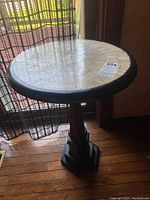 Round pedestal side table placed near a window with sheer curtains, showcasing the mosaic tile top and black border.