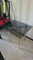 Clear square glass top side table with four chrome finish metal legs on carpeted floor beside black wooden chair with red cushion.