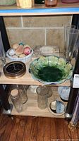Full shelf view showing marble eggs in a bowl, green ceramic bowl, square pink marble ashtray, two tall glass vases, and glass pitcher.