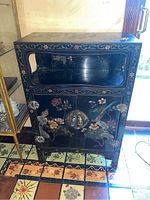 Front view of the black lacquered wood cabinet showing floral and bird painted motifs, open shelf, metal lock and latch on doors.