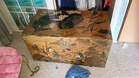 Full view of the vintage wooden chest with painted top surface showing lotus leaves and birds and sides with horseback riders and scenery.
