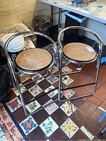 Pair of vintage chrome bar stools with curved back and caned seats on tiled floor, one needing repair.
