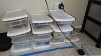 Stack of several clear plastic storage boxes with white lids arranged on the floor next to a black shelving unit.