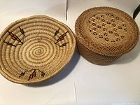 Two baskets shown: one open round basket with black and reddish-brown woven pattern, one round basket with lid featuring lattice woven design