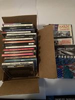 Top-down photo showing a box filled with multiple CDs and cassette cases, titles mostly related to 50's and 60's rock and roll music.
