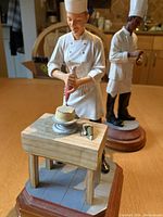 Pastry chef figurine decorating a cake with detail on wooden table and separate sous chef figurine in background.