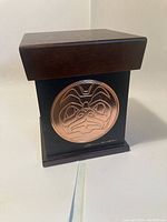 Front view of decorative box showing copper plate with First Nations art and artist's signature on black painted wooden box topped with darker wood lid.