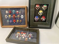 Overall view of three framed shadow box collections of miniature Asian masks on a table against a wall.
