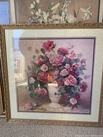 Large framed floral print showing a detailed vase of pink and white roses with a cream mat and gold frame placed up against another floral print.