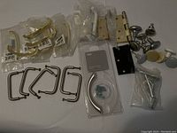 Photo showing multiple gold-tone cabinet handles still in packaging, metal rectangular pull handles, a gold-tone door hinge, metal and white knob pulls, and bags of screws and hardware.