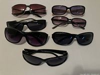Seven pairs of ladies sunglasses laid out on a white surface showing variety of frame styles and lens colors, mostly black frames with dark lenses.