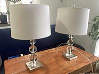 Two crystal glass ball table lamps with white drum shades on wooden table, shown from front angle.