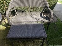 Front view of gray woven wicker love seat with matching ottoman on grass outdoors.